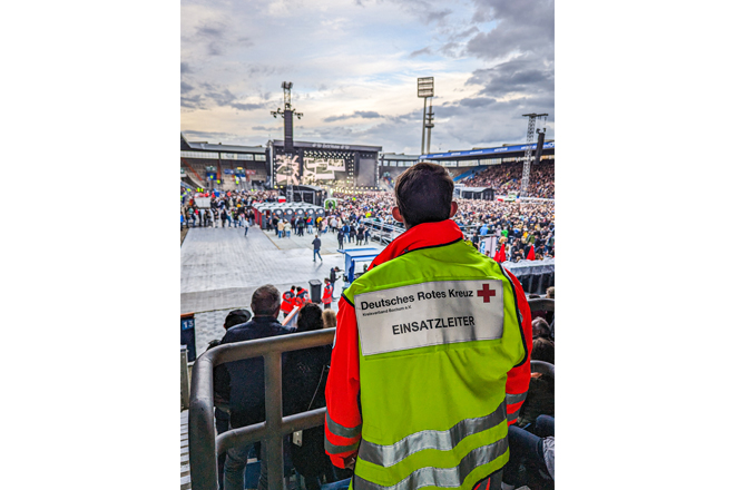 Mann mit DRK-Weste "Einsatzleitung" blickt auf ein Konzert im Stadion.