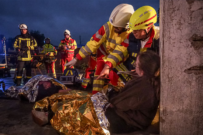 Zwei Sanitäter beraten sich neben einer an einer Säule gelehnten Frau mit Notfalldecke.