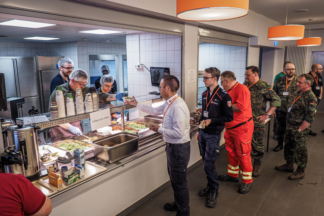 Zwei Mitarbeiter des DRK geben in einer Mensa Essen an wartende Menschen aus.