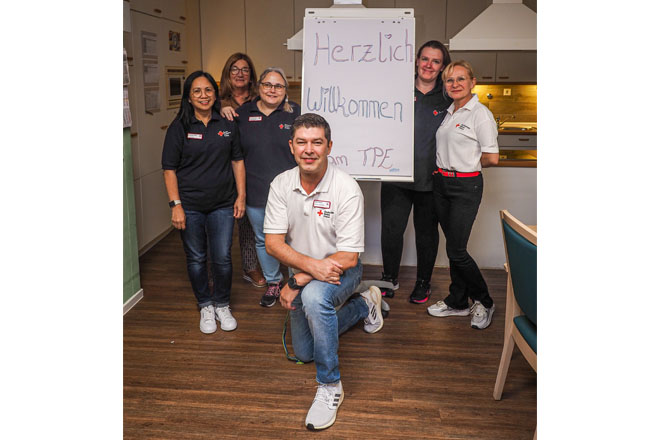 Sechs Menschen stehen um ein Schild "Herzlich Willkommen!". Der vordere Mann kniet.
