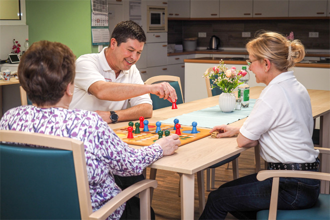 Zwei Pflegekräfte spielen mit einer Seniorin "Mensch ärgere Dich nicht".